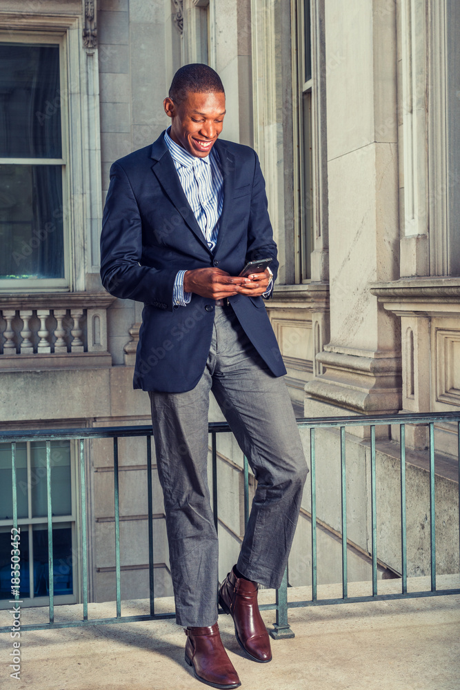 African American businessman in New York. Wearing black blazer, gray pants, brown leather shoes, a young guy standing in vintage style office, looking down, smiling, reading messages on mobile phone.. Stock Photo | African American businessman in New York. Wearing black blazer, gray pants, brown leather shoes, a young guy standing in vintage style office, looking down, smiling, reading messages on mobile phone.. Stock Photo |