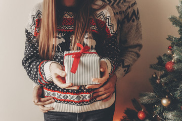 merry christmas and happy new year concept. stylish hipster couple in sweaters holding gift with red bow in festive room at christmas tree with lights. happy holidays. family  moments