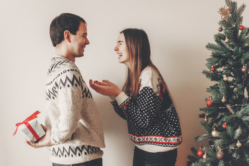 happy couple in stylish sweaters exchanging gifts in festive room at christmas tree. man holding surprise present behind back. merry christmas and happy new year concept