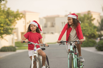 Two cute biracial kid sisters or friends wearing Santa hats while riding their bikes on a street in a neighbourhood with homes in the background