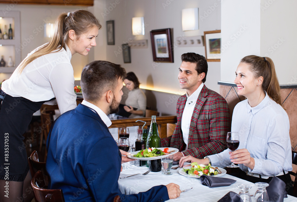 Polite smiling waitress bringing ordered dishes to guests at restaurant - obrazy, fototapety, plakaty 