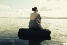 Rear View Of Woman Sitting On Rock In River Against Cloudy Sky