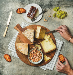 Poster - Flat-lay of cheese platter with cheese assortment, green grapes, honey and nuts with female hands reaching to food over grey concrete background, top view. Party or gathering eating concept