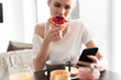 © Drobot Dean - Focused blonde lady eating bread with jam amd using smartphone
