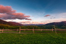 Wooden Fence On A Grassy Hill At Sunset. Beautiful Rural Scenery With Reddish Clouds Over The Mountain Ridge