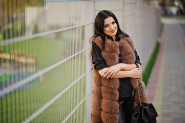 Fashion outdoor photo of gorgeous sensual woman with dark hair in elegant clothes and luxurious fur coat and with backpack against iron fence.