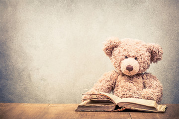 Wall Mural - Retro Teddy Bear toy sitting at the old wooden desk with old book front concrete wall background. Vintage instagram style filtered photo