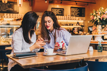 Two young happy women are sitting in cafe at table in front of laptop, using smartphone and laughing. On table paper notebook and cup of coffee. Girls are blogging, working, studying, learning online.