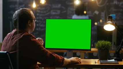 Office Employee at His Desktop Works on a Mock-up Green Screen Personal Computer. Over the Shoulder Footage. His Colleague works in the Background. Creative Office Evening. RED EPIC-W 8K Camera.