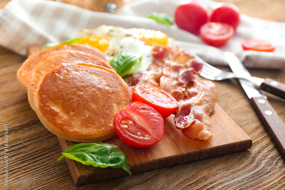 Board with yummy pancakes, tomatoes and fried bacon on wooden table