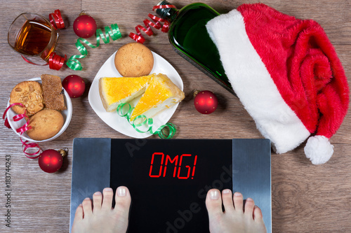 Fotografia Female feet on digital scales with sign "omg!" surrounded by Christmas decorations, bottle, glass of alcohol and sweets