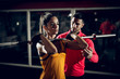 Close up motivated focused attractive young fitness woman doing squad exercise with a bar in front on the shoulders in the gym while her personal trainer standing next to her.