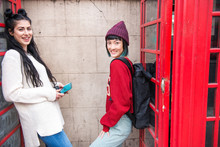 Portrait Of Two Young Stylish Women Leaning Against Red Phone Boxes, London, UK