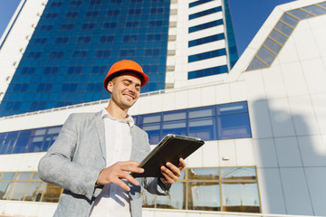 Successful business man in gray suit and protective construction orange helmet with tablet on background of building in blue white color. Engineer in helmet for workers security on office buildings.