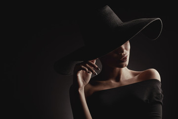 dramatic dark studio portrait of elegant woman in black wide hat and black dress. hidden eyes.