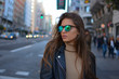 © franciscopgr - Mujer  joven estilosa  con gafas de sol y chaqueta negra caminando o realizando compras por la calle gran via de la ciudad de Madrid durante el atardecer