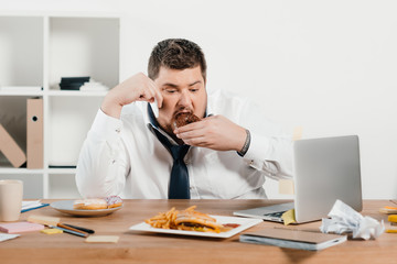 Wall Mural - overweight businessman eating donuts, hamburger and french fries at workplace