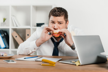 Wall Mural - overweight businessman eating donuts at workplace with laptop