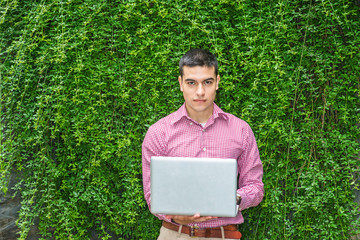 Wall Mural - College student studying in peaceful, green environment. Wearing red patterned, long sleeve shirt,  man standing against wall with long green leaves on campus, reading, working on laptop computer..