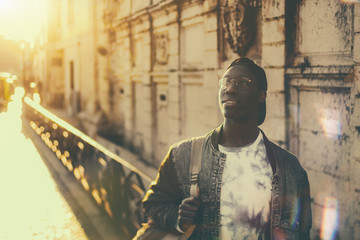 Black pensive undergraduate guy in denim jacket is standing on a street and looking up; handsome African male student nerd outdoor on a street of Lisbon city on his way to college or university