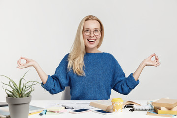 Positive carefree female office worker tries to relax after long hours of work, sits in lotus pose, meditates and concentrates, has happy expression, isolated over white background. Relaxed woman