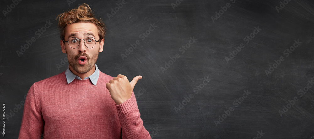 Horizontal shot of shocked male student with stylish hairdo, stares through spectacles, being surprised to indicate something at copy space for your promotional text or hearder. Advertising concept - obrazy, fototapety, plakaty 
