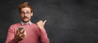 © WHstudio Leushin N - Fashionable male student holds modern smart phone, waits for call from groupmate, poses against black chalk board, indicates with thumb at copy space. Clever professor with electronic gadget.