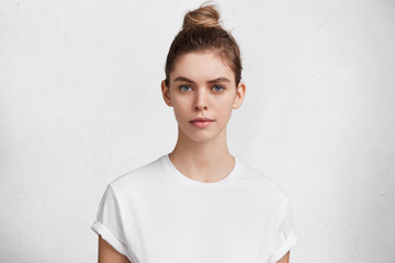 Serious pleasant looking woman with dark blonde hair tied in bun, wears casual white t shirt going to have walk outdoor with pet, has healthy pure skin. Pretty young female model poses in studio