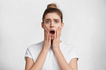 Wall Mural - Shocked female model with blue eyes keeps mouth widely opened, looks witth terrified expression, being afraid of awful things, isolated over white studio background. Pretty woman expresses surprisment