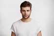 Serious confident young male student comes home after classes, dressed casually in white t shirt, waits for girlfriend, isolated over white background. People, youth, beauty, lifestyle concept