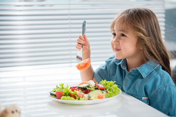 Side view profile of cute female child dreaming while eating salad in kitchen. Copy space