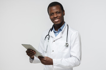 Wall Mural - Handsome african doctor standing isolated in grey background holding tablet device, showing confidence and optimism while giving straight open look and smiling