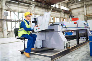 Concentrated operator wearing reflective jacket and hardhat focused on work while sitting in front of CNC machine, interior of spacious production department on background