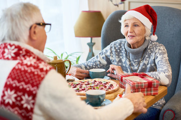 Poster - Portrait of loving senior couple celebrating Christmas together sitting at dinner table enjoying meal and talking, focus on elderly  woman wearing Santa hat