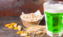 Glass With Green Beer And A Head Of Foam Near Plate With Pistachios, Wheat, Scattered Small Pretzels And Peanuts On Dark Desk
