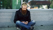 © silverkblack - Young bearded hipster man with headphones sitting on road and using smartphone for listen to music and internet surfing outdoors at street