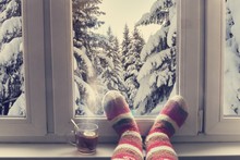 Feet In Wool Knitted Socks And A Cup Of Steaming Tea On The Windowsill Overlooking The Winter Snow Forest Through The Window