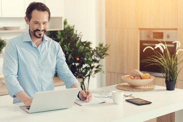 Wall Mural - Not to forget. Delighted happy positive man standing near the laptop and looking at its screen while taking notes