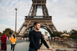 © Aleksandr - A young man wearing a dark blue jacket is standing on the background of the Eiffel Tower. Sunny weather is autumn