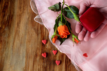 Male Hand Is Holding A Box For A Ring. An Orange Rose And Little Hearts Are Lying Next To It