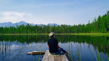 Lonely Dreaming Woman Sitting On A Wooden Bridge Pier Looking At The Mountains Reflected In The Mirror Water Of The Forest Lake. Serenity Concept. Pure Nature.