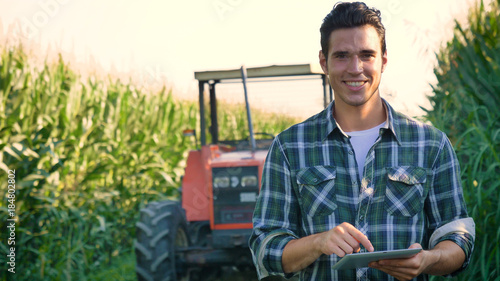 Fotomural Portrait of a beautiful young farmer (student) working in the field with a tractor working in a tablet, happy, in a shirt, corn field