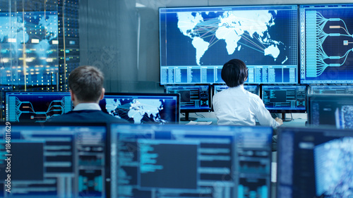 Fotomural In the System Control Room Operator and Administrator Sitting at Their Workstations with Multiple Displays Showing Graphics and Logistics Information