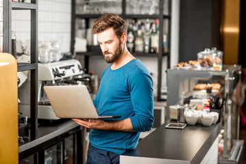 Handsome man in blue sweater working with laptop at the bar of the modern cafe interior