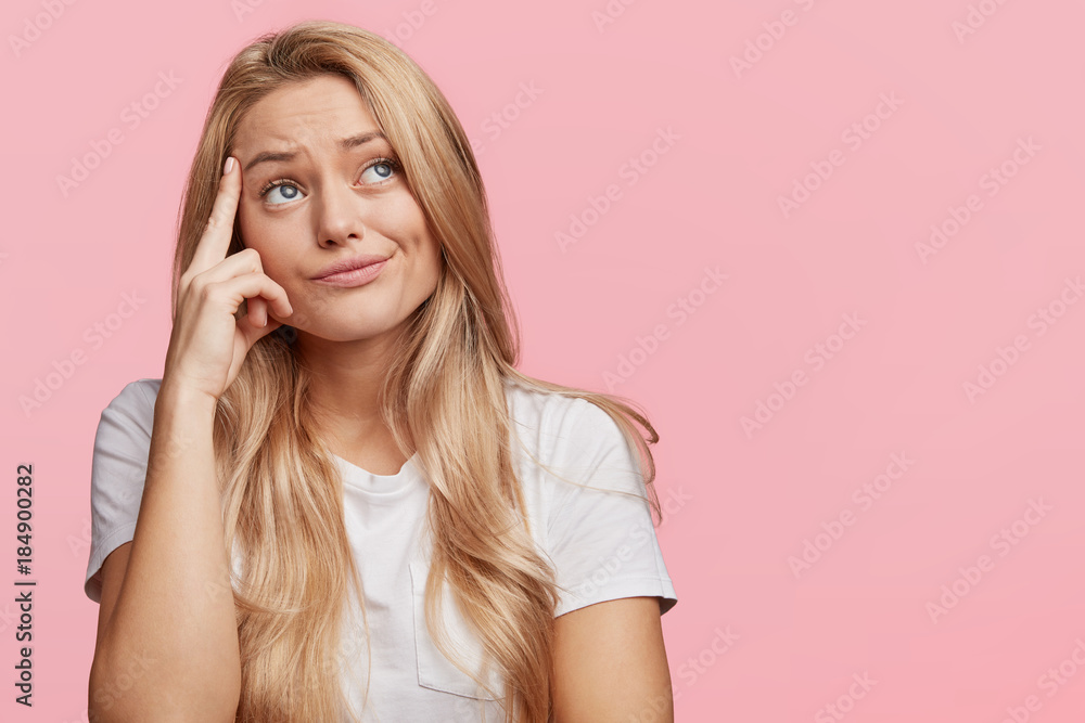 Photo of thoughtful young female with blonde hair and blue eyes, keeps finger on temple as tries to solve her serious problem, being deep in thoughts, poses against pink background with copy space - obrazy, fototapety, plakaty 