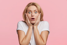 Scared European Woman Looks With Blue Bugged Eyes At Camera, Being Afraid Of Something, Keeps Hands On Cheeks, Looks In Terror, Isolated Over Pink Background. Emotional Horrified Female Indoor