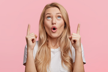 Wall Mural - Good looking blonde female with pure healthy skin looks in amazement as indicates at something upwards, isolated over pink background. Pretty young woman sees amazing thing up, gestures indoor