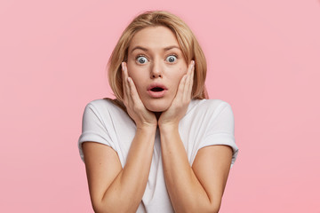 Wall Mural - Scared European woman looks with blue bugged eyes at camera, being afraid of something, keeps hands on cheeks, looks in terror, isolated over pink background. Emotional horrified female indoor
