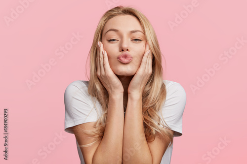 Portrait of lovely blonde female rounds lips as going to recieve kiss from boyfriend, looks at camera with pleased expression, isolated over pink background Tableau sur Toile