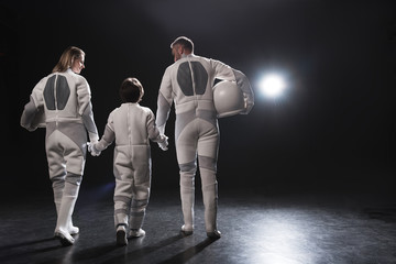 Friendly relations. Back view of positive family of astronauts are going together while holding by hands. Mother and father are looking at their little son. Dark background with copy space